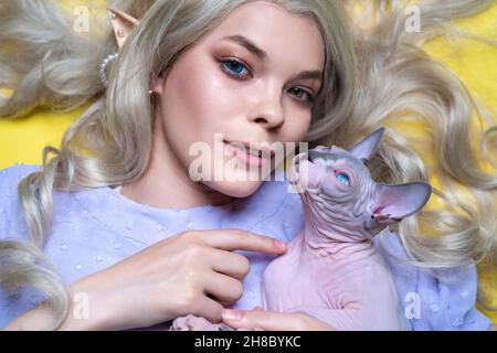 Porträt von Cosplay Elfe junge Frau in blauem Kleid, Luxus lockig blonde lange Haare, verschiedene Farben Augen, durchbohrte Ohren. Sie zeigt mit dem Finger auf die Sphinx-Katze Stockfoto