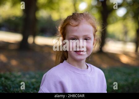Glückliches kleines Mädchen mit Sommersprossen und roten Haaren, das im Park die Kamera anschaut und lächelt. Stockfoto
