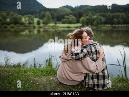 Rückansicht einer glücklichen älteren Mutter, die sich mit einer erwachsenen Tochter umarmt, wenn sie am See in der Natur sitzt Stockfoto