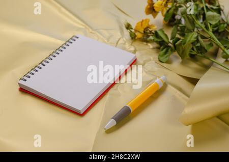 Öffnen Sie den federbelasteten Notizblock mit Stift auf cremefarbener Oberfläche. Verblassende gelbe Rosen im Hintergrund. Selektiver Fokus. Stockfoto
