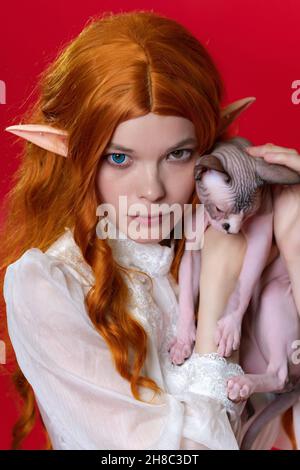 Porträt einer jungen Ingwer-Frau Cosplayer Elf hält in den Händen Sphinx Kätzchen, die mit seinen Pfoten, Schwanz hängt nach unten, schlampt. Elfe in weißem Kleid Stockfoto