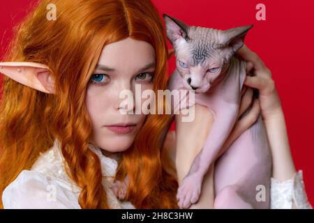 Porträt einer jungen Cosplayerin-Elfe mit reinrassigen Sphinx-Kätzchen mit blauen Augen. Ingwer-Elf im Sommer weißen Kleid, mit langen Haaren, hält Kätzchen Stockfoto