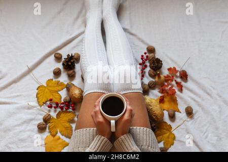 Weibliche Hände halten eine Kaffeetasse auf den Beinen in wollenen Wintersocken in einem gemütlichen Bett mit Herbstblättern zu Hause dekoriert. Nahaufnahme, selektiver Fokus Stockfoto