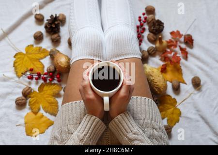 Frau Hände halten eine Tasse Kaffee auf den Beinen in wollenen Wintersocken in einem gemütlichen Bett mit Herbstblättern zu Hause dekoriert. Nahaufnahme, selektiver Fokus Stockfoto