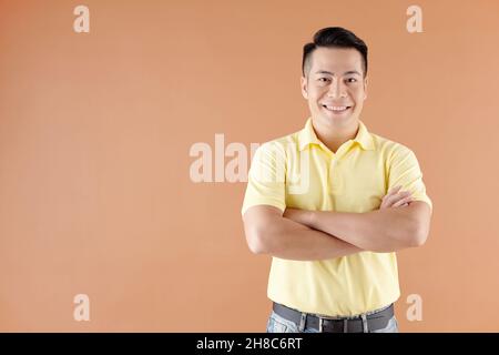 Porträt eines lächelnden jungen asiatischen Mannes, der mit gekreuzten Armen vor braunem Hintergrund steht Stockfoto