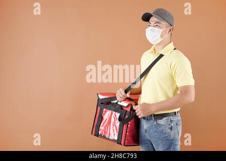 Porträt der Lieferung Mann in medizinische Maske hält Tasche mit Pizza und wegschauen Stockfoto