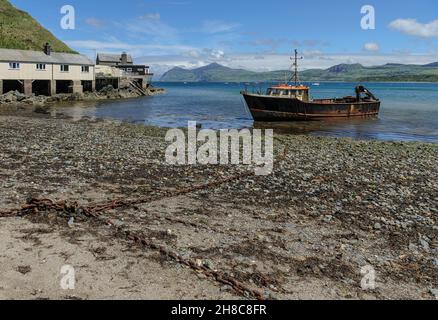 An einem hellen Sommertag mit blauem Himmel und leichten Wolken wird das Boot von Ketten zum Kies- und Kiesstrand mit freiem Blick über die Bucht zu den fernen Hügeln vertäut. Stockfoto