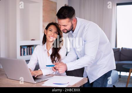 Glückliches Paar Online-Shopping von zu Hause aus. Ein hübscher Mann hält eine Bankkarte in der Hand, während ihre Frau neben ihm sitzt und beim Einkaufen einen Laptop benutzt Stockfoto