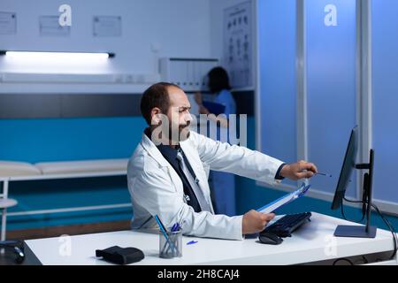 Konzentrierter Arzt, der bei Nachtschicht auf den Monitor zeigt, um Untersuchungen durchzuführen. Arzt, der auf den Computerbildschirm schaut, um Analyseinformationen und medizinische Versorgung zu erhalten. Facharzt, der spät arbeitet Stockfoto