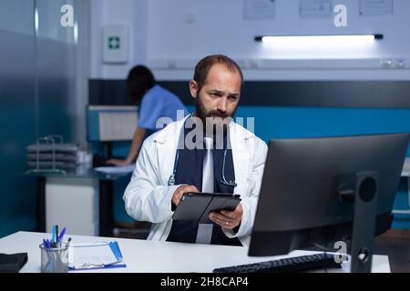Spezialist, der Technologie für Gesundheitssysteme und Praxis im Büro verwendet. Arzt hält ein digitales Tablet, während er auf den Computerbildschirm schaut, um Informationen zur Analyse zu erhalten, und arbeitet spät. Stockfoto