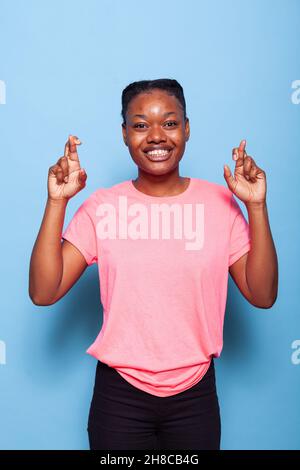 Porträt eines lächelnden afroamerikanischen Studenten mit gekreuzten Fingern, das das Symbol des Begehrens im Studio mit blauem Hintergrund zeigt. Junge Frau, die für Viel Glück. betet Cross-Finger-Konzept Stockfoto