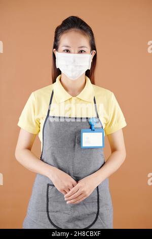 Porträt einer positiven jungen Kellnerin, die mit einer medizinischen Maske auf ihrem Gesicht steht und die Kamera anschaut Stockfoto