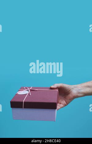 Goiânia, Goias, Brasilien – 29. November 2021: Die Hand älterer Menschen bietet eine Geschenkbox mit blauem Hintergrund an. Stockfoto