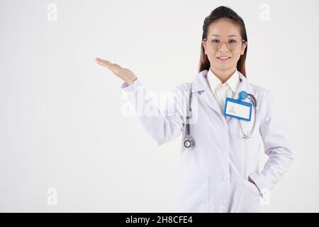 Lächelnde, hübsche junge Ärztin mit Abzeichen auf der Brust, die etwas mit der Hand zeigen Stockfoto