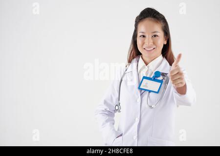 Fröhliche, hübsche junge Praktizierende lächelt und zeigt den Daumen nach oben Stockfoto