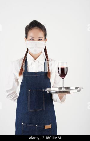 Porträt einer positiven jungen Kellnerin in Schutzmaske mit Tablett mit Glas Rotwein Stockfoto