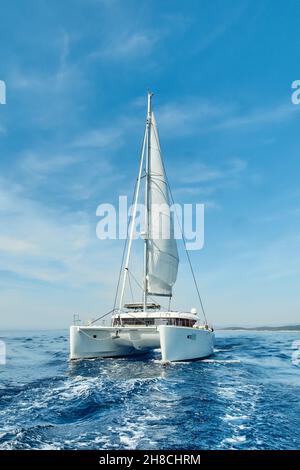 An einem sonnigen Sommertag segelt ein luxuriöser Katamaran unter weißen Segeln auf dem blauen azurblauen Meer. Reisen, Segeln, Regatten Konzept Stockfoto