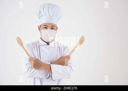 Portrait des fröhlichen Küchenchefs in medizinischer Maske mit Holzlöffel und Spachtel Stockfoto
