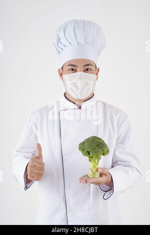 Portrait eines positiven Küchenchefs in Schutzmaske mit frischem Brokkoli und Daumen nach oben Stockfoto