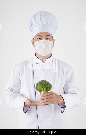 Portrait eines selbstbewussten Küchenchefs mit Hut, weißer Uniform und medizinischer Maske mit frischem Brokkoli Stockfoto