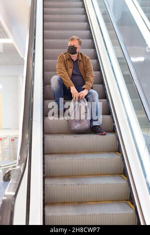 Ein müder Erwachsener in einer Schutzmaske mit Rucksack reitet auf einem Excalator in der U-Bahn sitzen. Stockfoto