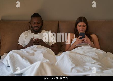 Lächelnder Mann und fokussierte Frau, die ihre Handys im Schlafzimmer benutzte Stockfoto