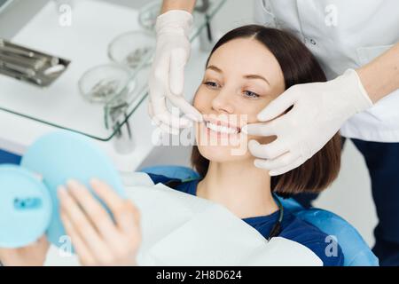 Schau mich an. Attraktive kaukasische Dame überprüft ihr schönes Lächeln im Spiegel nach stomatologischer Behandlung. Zufriedenheit des Besuches Stomatologen Stockfoto