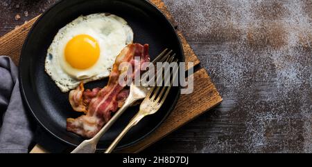Traditionelles englisches Frühstück mit Spiegeleiern und Speck in einer gusseisernen Pfanne auf dunklem Betonboden. Draufsicht. Stockfoto