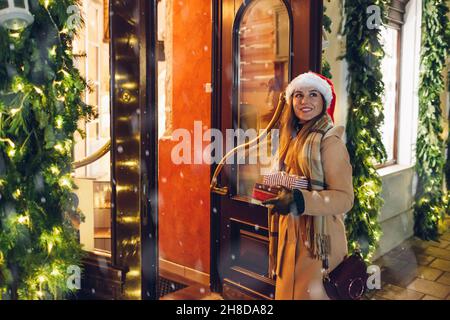 Happy Woman in Santa hat Shopping für Weihnachtsgeschenke hält Geschenkboxen Betreten dekorierten Laden auf der Straße der Stadt Stockfoto