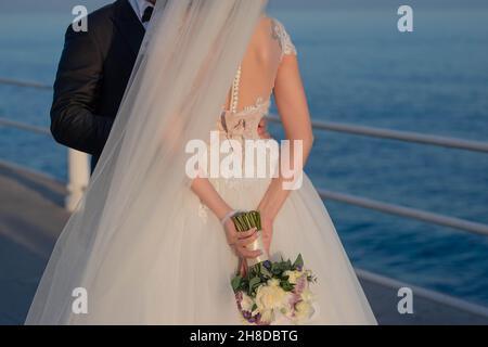 Schöne Braut und Bräutigam in einer romantischen Umarmung, die Frau trägt ein exquisites Hochzeitskleid und hält einen eleganten Blumenstrauß auf dem Rücken Stockfoto