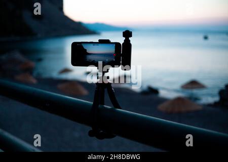 Mobiltelefon mit tragbarer Kamera, die auf einem Stativ montiert ist und den Sonnenaufgang über der Meeresbucht filmt. Videoaufnahme eines Zeitrahals des Sonnenuntergangs über Felsen im Wasser mit mobilem Pho Stockfoto