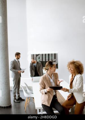 Zwei junge Geschäftsfrauen, die im Büro mit jungen Menschen über digitale Tablets diskutieren, arbeiten hinter ihnen Stockfoto