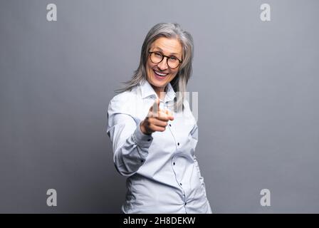 Hey, du. Lächelnd positiv glücklich Senior grauhaarige Geschäftsfrau schaut auf die Kamera und zeigt Zeigefinger auf Sie, reife Dame in formeller Kleidung stehend isoliert auf grau, wählt Sie aus Stockfoto