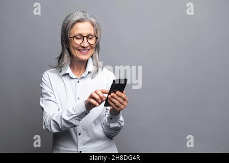 Fröhliche positive glückliche ältere grauhaarige Geschäftsfrau, die ein Smartphone benutzt, das isoliert auf einer grauen, reifen Dame in formeller Kleidung steht und Touchscreen, scrollende Neuigkeiten, Nachrichten, SMS-Nachrichten anfasst Stockfoto