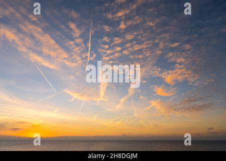 Kondensstreifen & leichte, flauschige Wolken über dem Meer, mit einer orangefarbenen Tönung, die vom morgendlichen Sonnenaufgang im Winter in Großbritannien erhellt wird. Wolken von der Sonne beleuchtet. Stockfoto