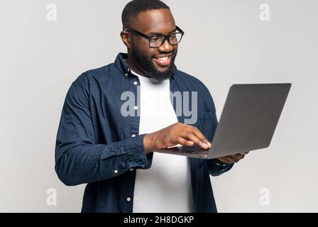 Ein kluger und konzentrierter afroamerikanischer Geschäftsmann, der ein legeres Hemd und eine Brille mit einem Laptop trägt, ein männlicher Büroangestellter, der Computer hält und eine Nachricht tippt, isoliert auf Grau Stockfoto