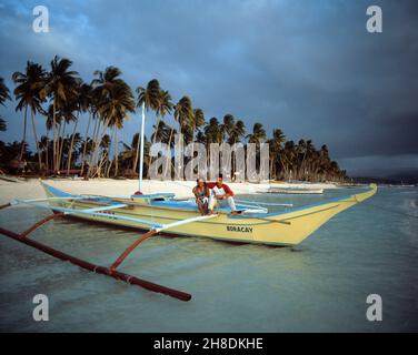 Philippinen. Boracay Island. Junges einheimisches Paar am Strand, das auf einem Auslegerkanu sitzt. Stockfoto