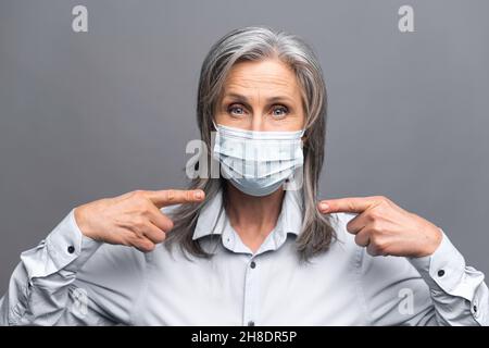 Seriöse ältere Frau in formeller Kleidung und medizinische Maske auf dem Gesicht zeigt Finger isoliert auf grauem, Nahaufnahme Porträt einer weiblichen Büroangestellte schützen sich vor Viruserkrankungen, empfiehlt Maßnahmen Stockfoto