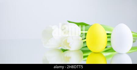 Hühnereier, weiß und gelb auf grauweißem Hintergrund gemalt, spiegeln sich auf der Spiegelfläche neben dem Bouquet mit weißen Tulpen. Kreati Stockfoto