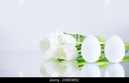 Hühnereier, weiß auf grauem Grund gemalt, spiegeln sich auf der Spiegelfläche neben dem Bouquet mit weißen Tulpen. Kreative Karte mit East Stockfoto