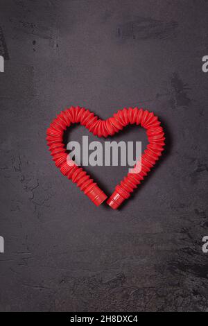 Gestreckt leuchtend rot Pop it tube Kinder Spielzeug gebogen in Form von Herz. Kunststoff-Anti-Stress-Spielzeug in der Mitte der schwarzen strukturierten Stein Hintergrund. Flach liegend. C Stockfoto