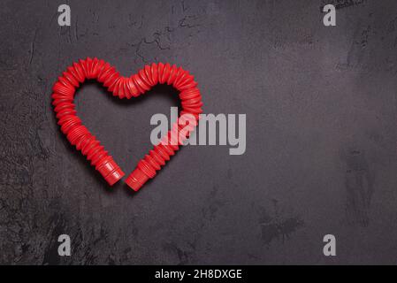 Hell gestreckt rot Pop it tube Kinder Spielzeug gebogen in Form von Herz. Kunststoff-Anti-Stress-Spielzeug auf schwarzem texturierten Stein Hintergrund. Flach liegend. Große Kopie Stockfoto