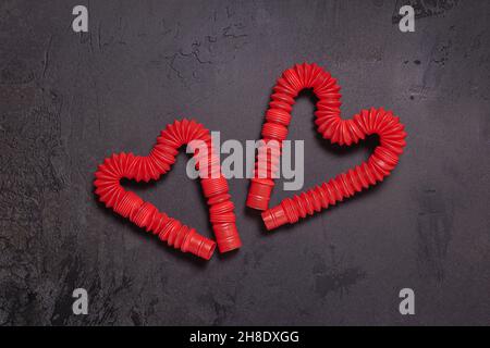 Zwei gestreckte rote Pop-it-Tube Kinder Spielzeug in Form von Herzen gebogen. Kunststoff-Antistress helle Spielzeug auf schwarzem texturierten Stein Hintergrund. Flach liegend. Kopieren Stockfoto