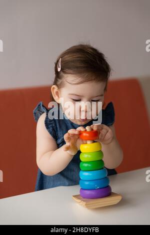 Ziemlich nettes Baby Mädchen trägt Jeans Kleid spielen bunte Spielzeug hölzerne Pyramide und machen Schießen emotionales Gesicht. Kleines Kind interessant erforscht und Stockfoto