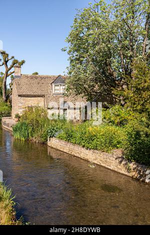 Ein traditionelles Steinhaus neben dem kleinen River Windrush, der durch das Cotswold-Dorf Naunton, Gloucestershire, Großbritannien, fließt Stockfoto