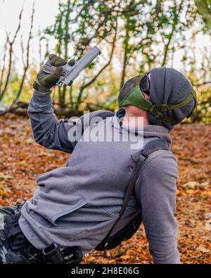 Airsoft-Spieler, der mitten in einem Spiel im Wald eine Pose für die Kamera aufwirft, hält eine benutzerdefinierte Handfeuerwaffe und trägt Schutzausrüstung Stockfoto