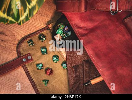 Overhead-Bild einer offenen braunen Lederkuriertasche mit einem Würfelbeutel mit Rollenspielwürfeln und einem Ledernotizbuch und einem Stift Stockfoto