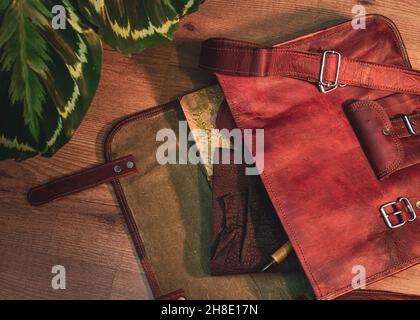 Überkopfbild einer offenen braunen Umhängetasche aus Leder mit einem Buch, einem Ledernotizbuch und einem Stift Stockfoto