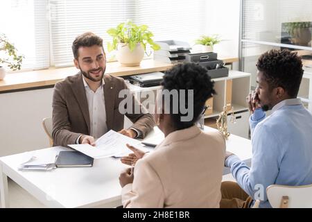 Lächelnd hübscher junger bärtiger Anwalt in Jacke, der am Tisch sitzt und dem Paar im Büro Dokumente gibt Stockfoto