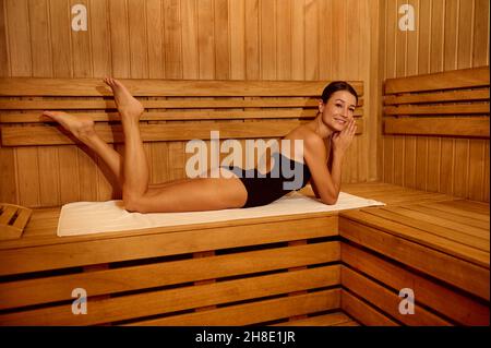 Attraktive Frau mittleren Alters in schwarzem Badeanzug, die sich in einer traditionellen finnischen Holzsauna entspannt, auf dem Bauch auf einer Bank liegt, scharfsinnig lächelt und schaut Stockfoto